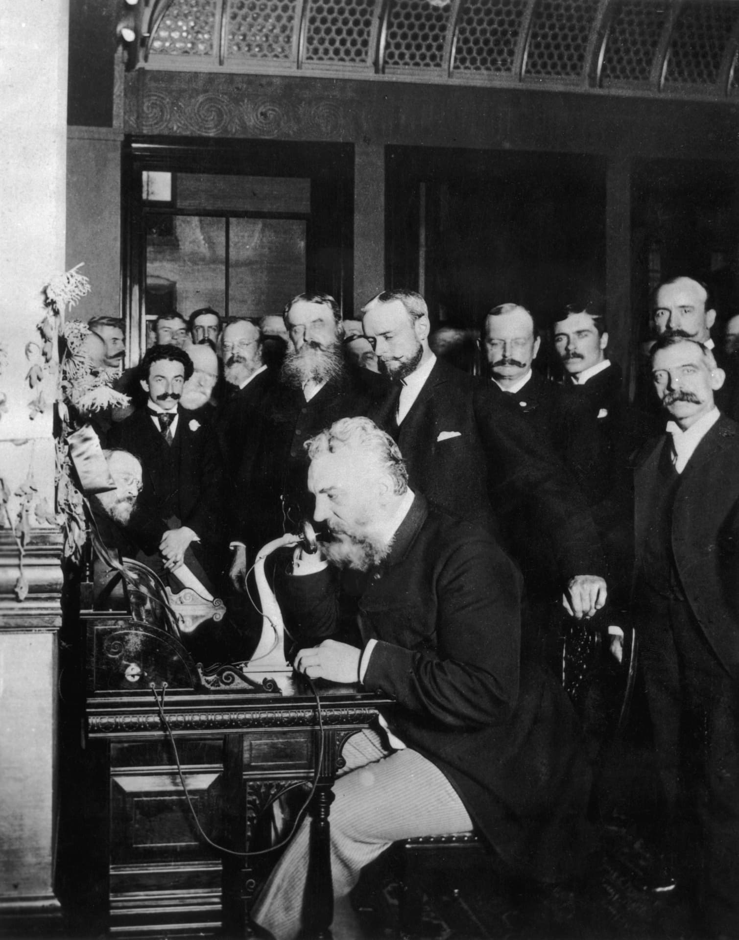 Historical black-and-white photograph of Alexander Graham Bell speaking into an early telephone, watched by a group of onlookers